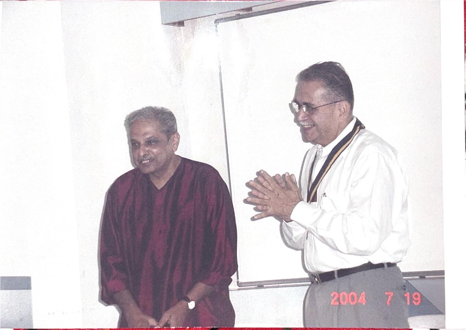 Two Rotarians standing before a whiteboard at a club meeting, one welcoming the other with folded hands.