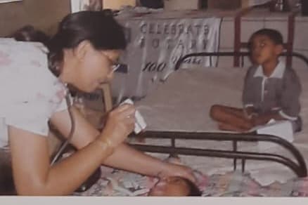 A health worker with a stethoscope examining a child on a cot at a community camp, a Celebrate Rotary banner behind her.